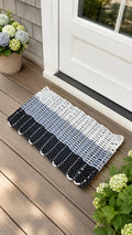 A woven outdoor doormat with a striped pattern in black, gray, and white, placed on a wooden deck in front of a white door with potted hydrangeas nearby.