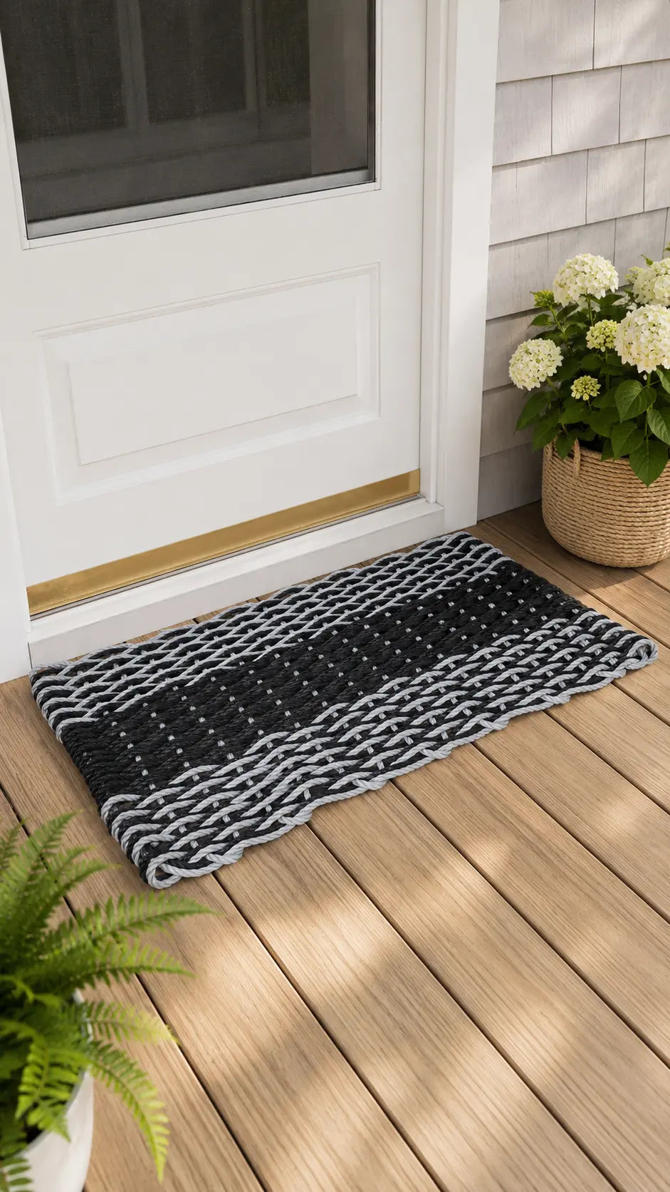 A black and gray braided doormat with a textured pattern, placed on a wooden deck in front of a white front door, with potted plants nearby.