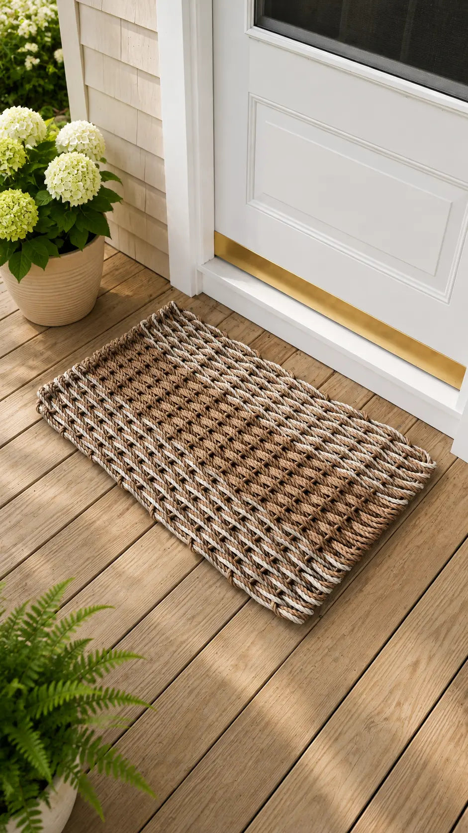 A rectangular woven doormat with a braided houndstooth-like pattern in natural tan, brown, and cream, placed on a wooden deck in front of a white front door.