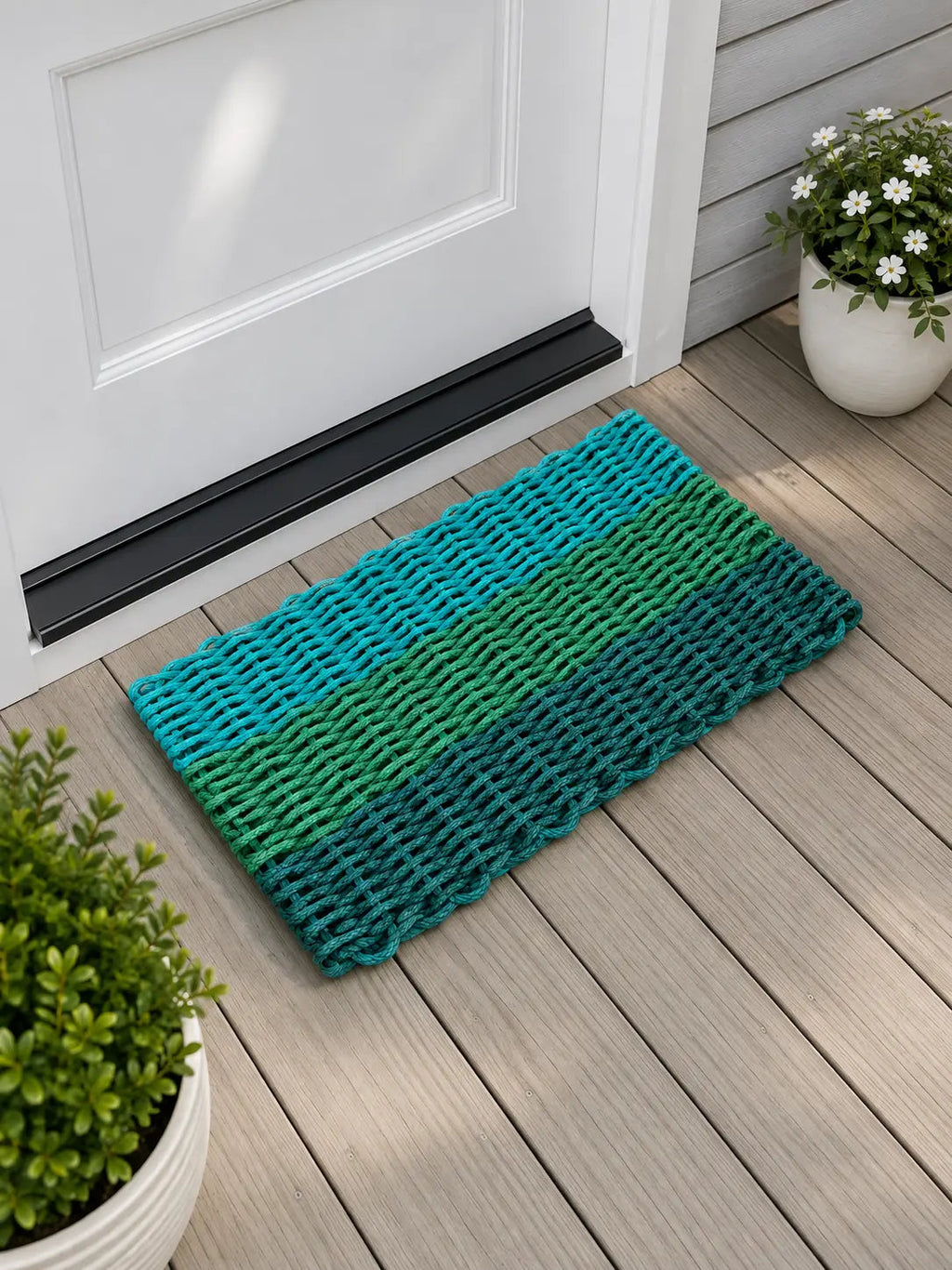 A rectangular woven doormat with horizontal stripes in teal, green, and dark blue-green, placed on a light wood deck in front of a white door with potted plants nearby.