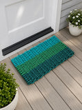 A rectangular woven doormat with horizontal stripes in teal, green, and dark blue-green, placed on a light wood deck in front of a white door with potted plants nearby.