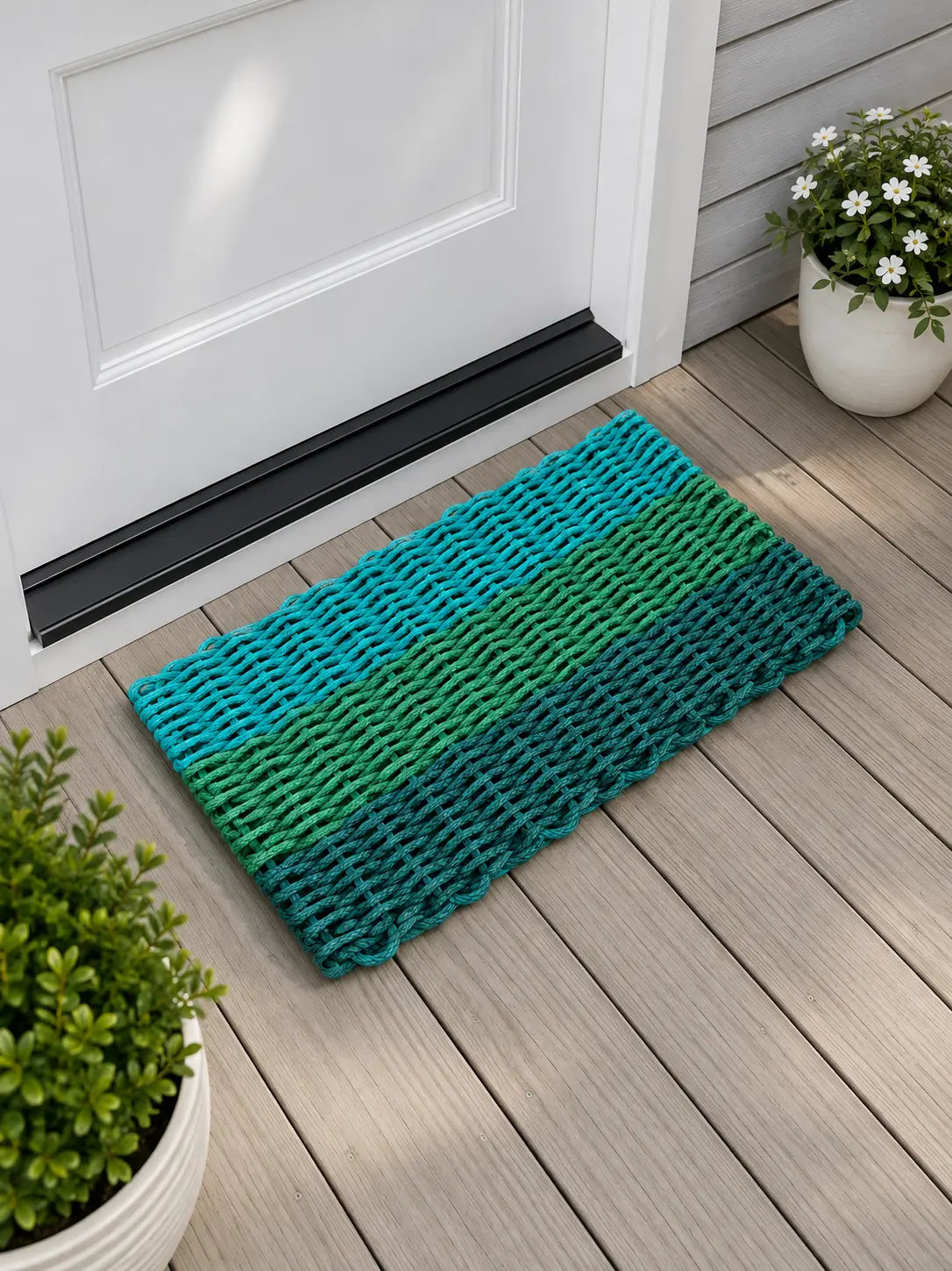 A rectangular woven doormat with horizontal stripes in teal, green, and dark blue-green, placed on a light wood deck in front of a white door with potted plants nearby.