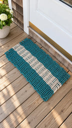 A rectangular woven doormat with teal blue and off-white horizontal stripes, placed on a wooden deck in front of a white door.