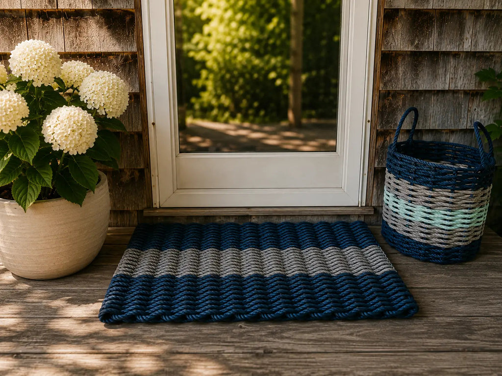 A rectangular woven doormat with horizontal navy, gray, and white stripes lies on a wooden porch floor in front of a white-framed glass door. A beige planter with white flowers sits on the left, and a blue-and-white basket is on the right.

