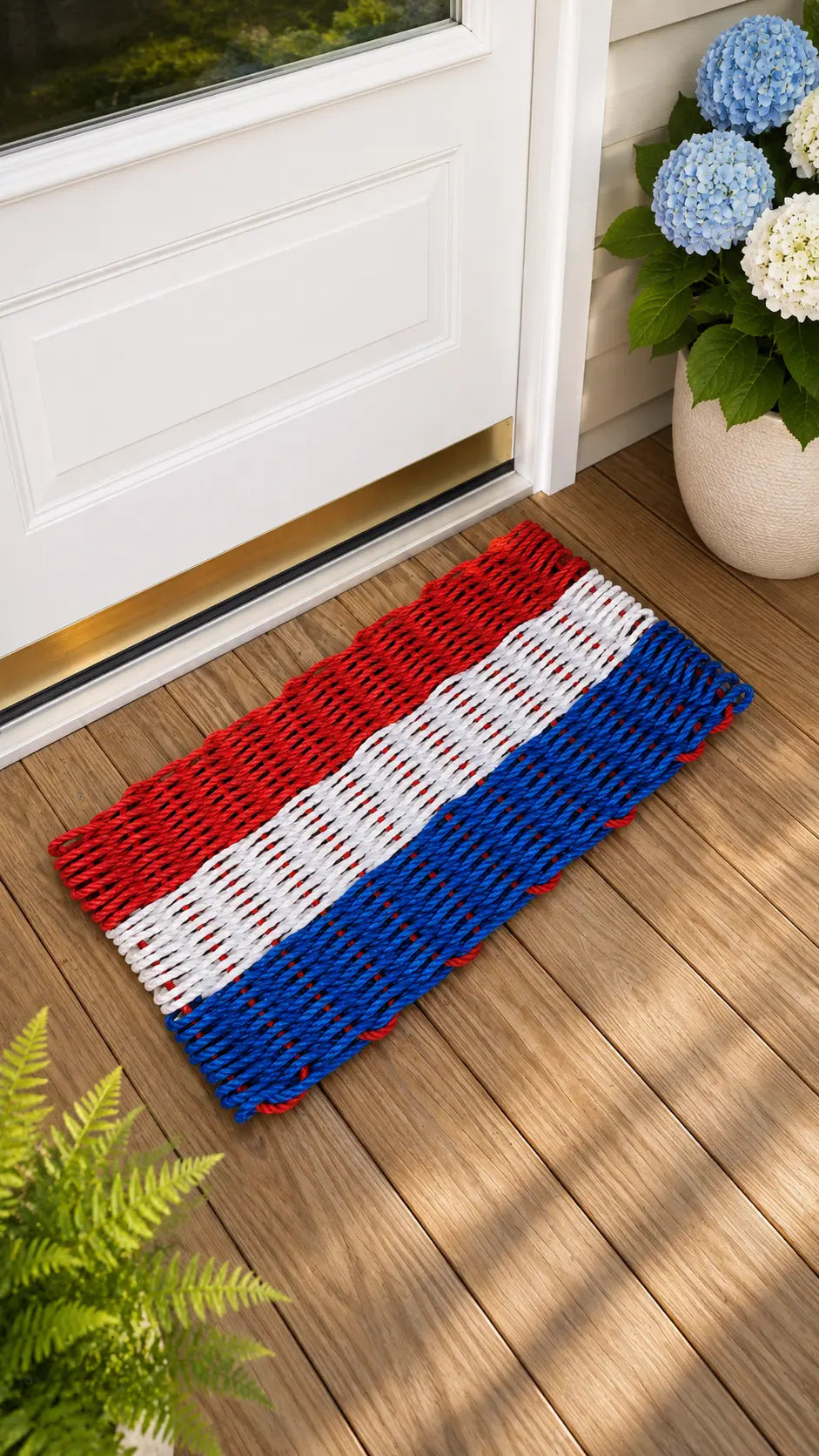 A woven doormat with horizontal red, white, and blue stripes lies on a wooden deck in front of a white door. A potted plant with blue and white flowers sits to the right, and green fern leaves appear in the lower left corner.