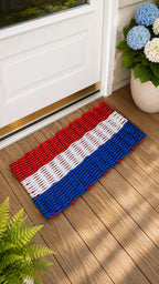 A woven doormat with horizontal red, white, and blue stripes lies on a wooden deck in front of a white door. A potted plant with blue and white flowers sits to the right, and green fern leaves appear in the lower left corner.