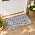 A rectangular woven doormat in a light gray color with a scalloped edge, placed on a wooden deck in front of a white door. A wicker basket with white flowers and a potted fern are visible in the background.
