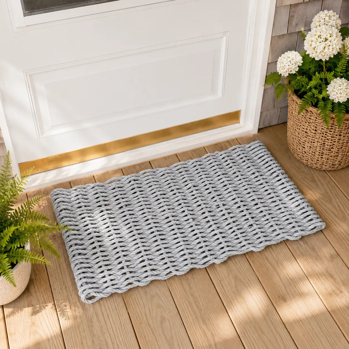 A rectangular woven doormat in a light gray color with a scalloped edge, placed on a wooden deck in front of a white door. A wicker basket with white flowers and a potted fern are visible in the background.