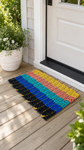 A colorful woven doormat with horizontal stripes in black, blue, teal, yellow, orange, and pink, placed on a wooden front porch next to a white door and potted plants.