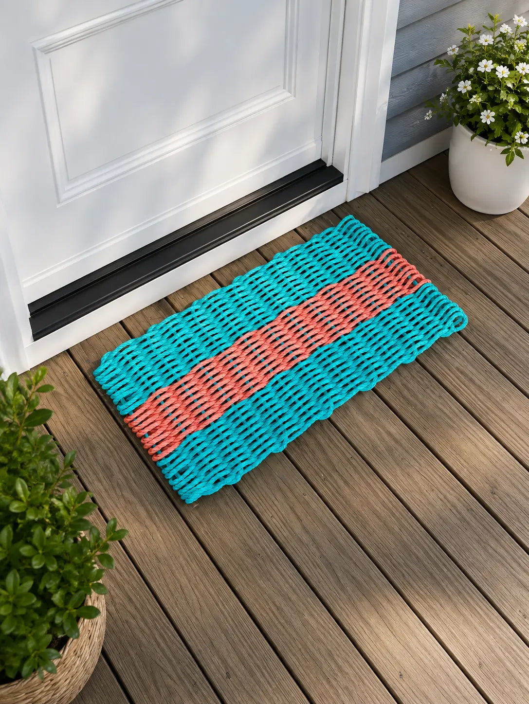 A rectangular woven doormat with turquoise, coral, and white horizontal stripes, placed on a brown wooden deck in front of a white door with potted plants nearby.