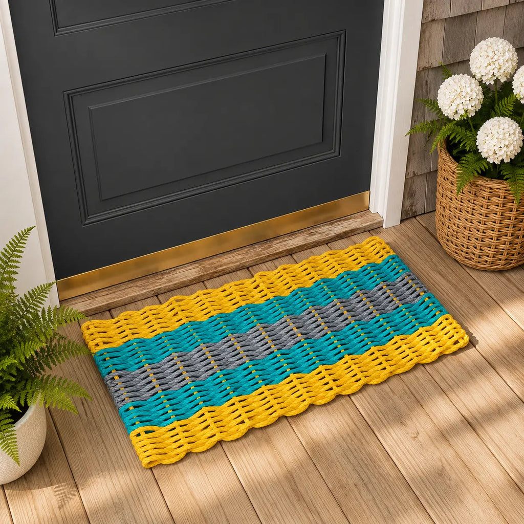 A woven rectangular doormat with yellow, teal, and gray horizontal stripes, placed in front of a dark charcoal door on a light wood floor, with potted plants nearby.