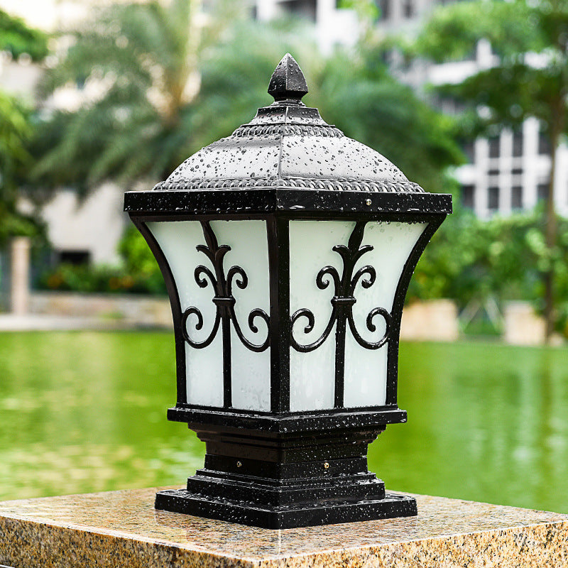 Black metal outdoor garden lamp with frosted white glass panels and ornate scrollwork, displayed on a stone pedestal near a pond and trees.