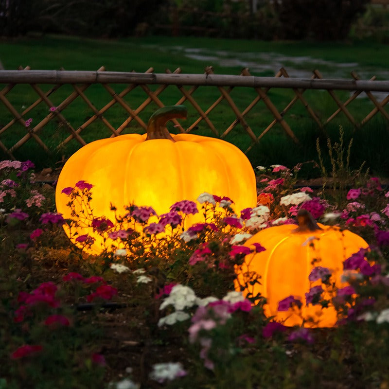 Two illuminated pumpkin decorations displayed in a garden with flowers and a wooden fence in the background.