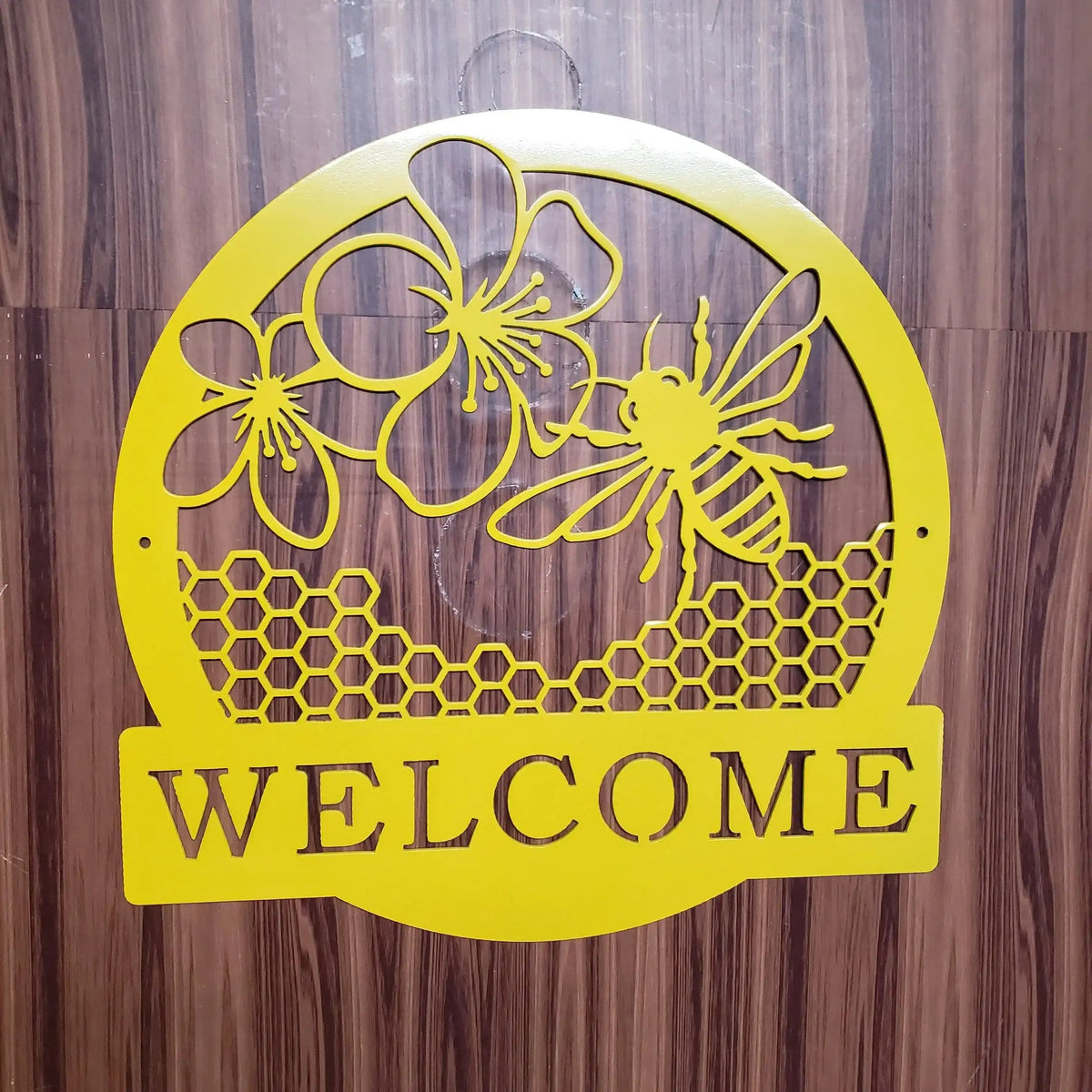 Yellow metal wall sign with a bee, flowers, honeycomb pattern, and the word 'WELCOME' in cutout letters, mounted against a dark wood-paneled wall.